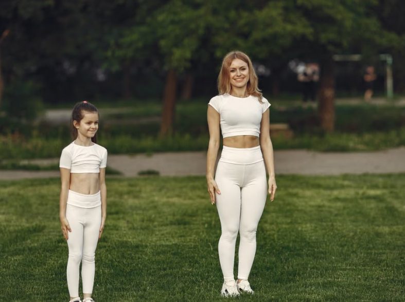 A woman and girl in white activewear standing on a grass field outdoors, smiling.
