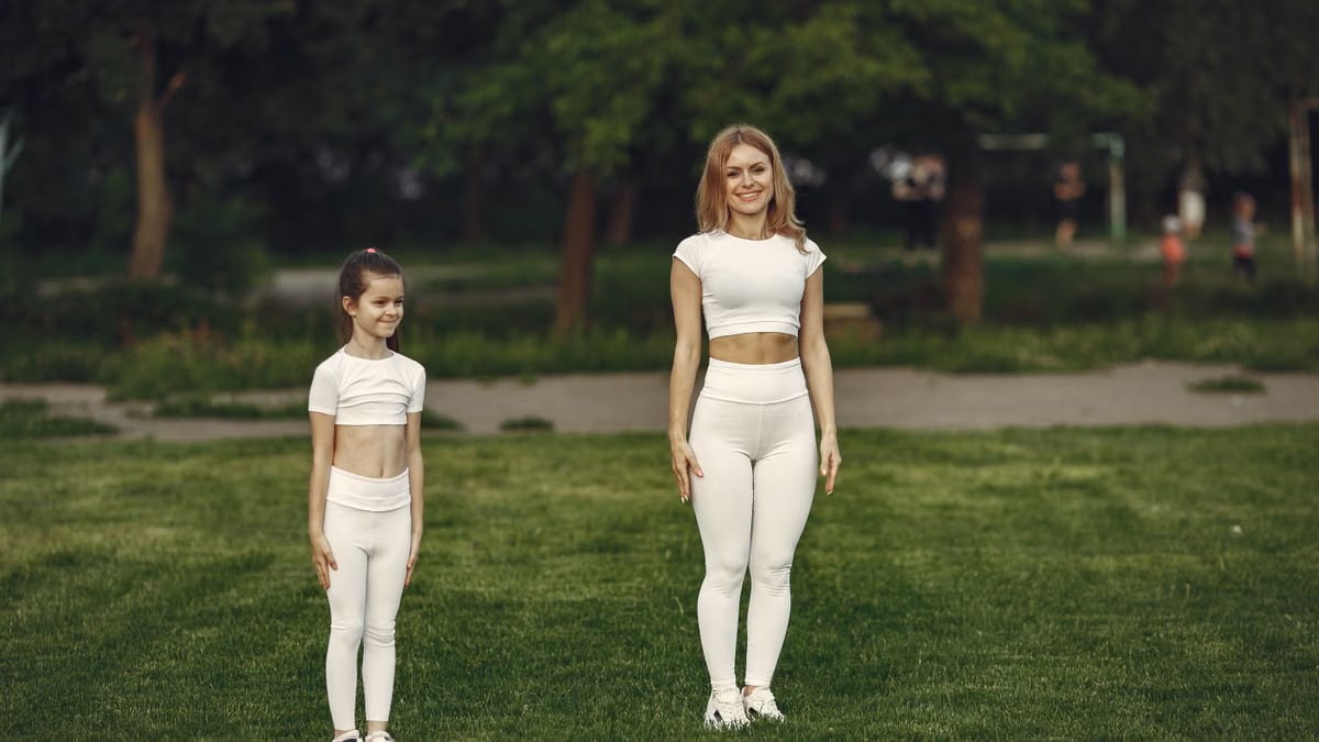 A woman and girl in white activewear standing on a grass field outdoors, smiling.